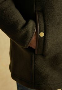 Dłoń częściowo w kieszeni ciemnozielonej polarowej kurtki z brązowymi wykończeniami i małym okrągłym złotym guzikem na krawędzi kieszeni.