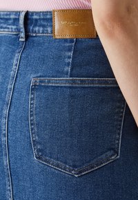 Denimhame, joka on keskisinisen sävyinen, takapockettiyksityiskohta, ruskea nahkaleima kohokuvioidulla tekstillä, sileä pinta ja klassinen istuvuus.