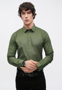 Olivově zelená košile na knoflíky s límcem, dlouhými rukávy a hladkou texturou; doplněná o černé hodinky a tmavé džíny.