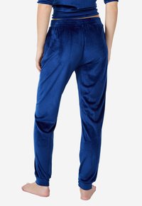 Osoba ubrana w niebieskie dresowe spodnie z weluru oraz pasujący długi top, stojąca boso tyłem na białym tle.