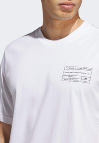 Hvid bomulds T-shirt med en sort trykt etiket på venstre bryst med tekst og et logo. Rund halsudskæring og korte ærmer.