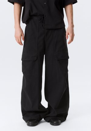 Vyras dėvi laisvus juodus krovinių kelnes su didelėmis priekinėmis kišenėmis ir juodus batus prieš paprastą foną.