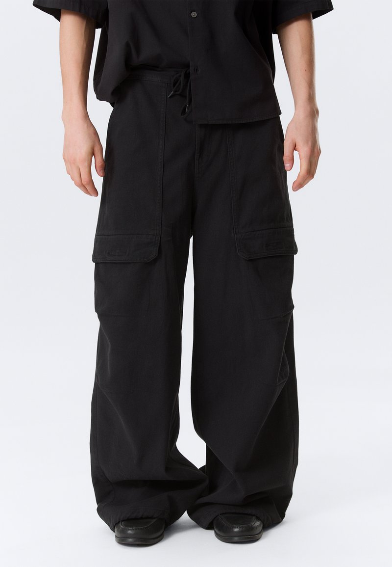 Άνδρας που φοράει φαρδιά μαύρη παντελόνα cargo με μεγάλες μπροστινές τσέπες και μαύρα παπούτσια, μπροστά σε απλό φόντο.