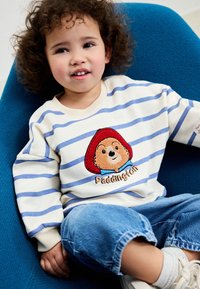 Sweat-shirt blanc avec des rayures bleues, arborant un motif brodé de l'ours Paddington à l'avant. Pantalon en denim décontracté et chaussures faciles à enfiler.