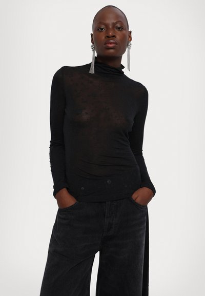 Top lung cu maneci lungi, de culoare neagră, dintr-un material transparent și texturat, cu guler tip mock; asortat cu pantaloni largi negri, cu mâinile în buzunare; având modele subtile.