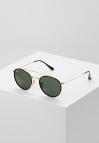 Ochelari de soare cu ramă din metal aurie, cu lentile ovale verzi, brațe subțiri, vârfuri maronii închise și un design minimalist.