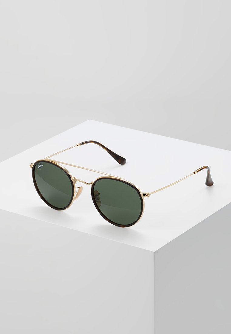 Ochelari de soare cu ramă din metal aurie, cu lentile ovale verzi, brațe subțiri, vârfuri maronii închise și un design minimalist.