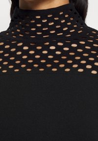 Haut noir à col montant avec un design de découpes circulaires sur toute la partie supérieure, contrastant avec un tissu uni en dessous.