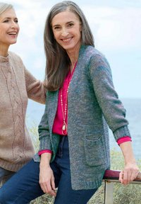 Deux femmes âgées souriant en plein air près de la mer, l'une portant un cardigan multicolore et un blouse rose, l'autre dans un pull beige en tricot.