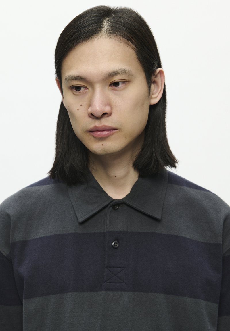 Mørk grå og navy stribet polo t-shirt med krave, med en knaplukning og tekstureret stof. Cirkulære sorte knapper synlige.
