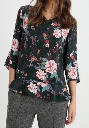 Vrouw draagt een donkergroene bloes met roze en witte bloemen en grijze gestructureerde broek, staand met één hand in de zak.