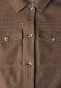 Brun ullskjorta med krage, som har två bröstfickor och stora knappar. Tyget har en mjuk, strukturerad yta.