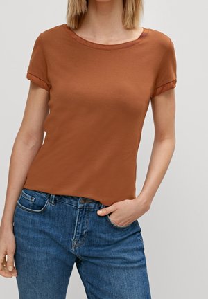 Femme portant un t-shirt marron ajusté à manches courtes et un jean bleu, une main dans la poche, sur fond uni.