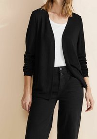 Svart stickad cardigan med öppen front, långa ärmar och ribbade manschetter. Kombinerad med en vit topp och svarta högmidjade byxor.