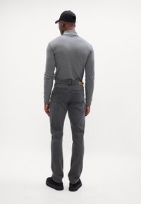 Maglione grigio a maniche lunghe e jeans grigi con tasche posteriori, indossato con scarpe nere e un cappellino nero, visto da dietro su uno sfondo chiaro.