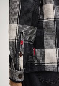Zwart-wit geblokte textuur jas mouw met bruine knopen en rode stoffen labels, gedragen over donkerdenim jeans.