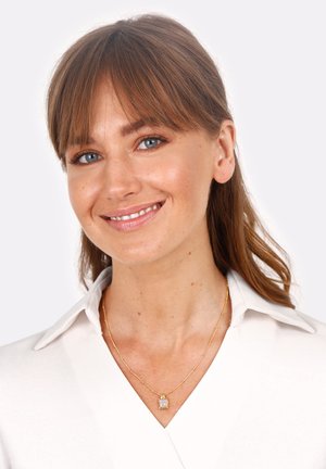 Goldene Halskette mit einem rechteckigen Anhänger, getragen über einem weißen Hemd mit Kragen. Frau lächelt mit braunen Haaren und blauen Augen.
