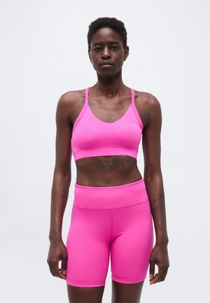 Femme debout, face à l'avant, portant un soutien-gorge de sport rose vif et un short de cycliste taille haute assorti, sur un fond blanc uni.