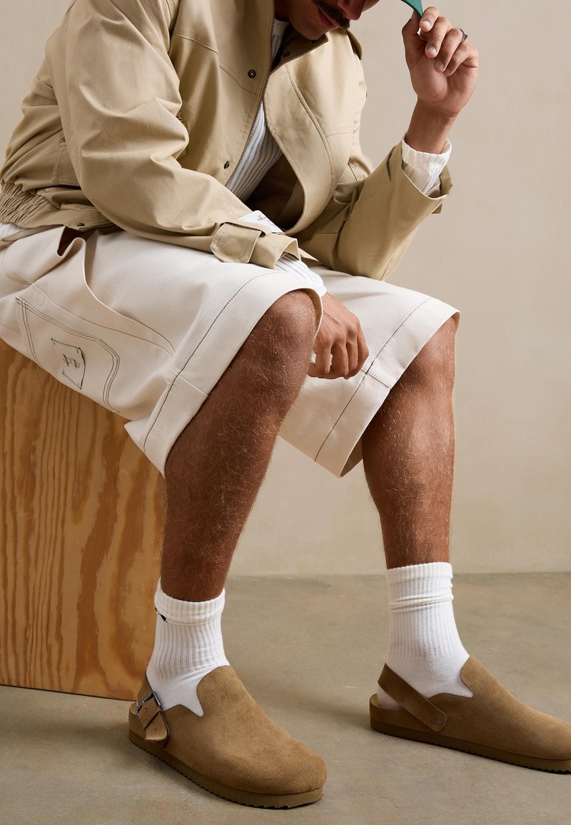 Homme assis sur un bloc de bois, portant une veste beige, un short blanc, des chaussettes blanches, des chaussures en daim marron clair sans lacets, et tenant une casquette verte devant son visage.