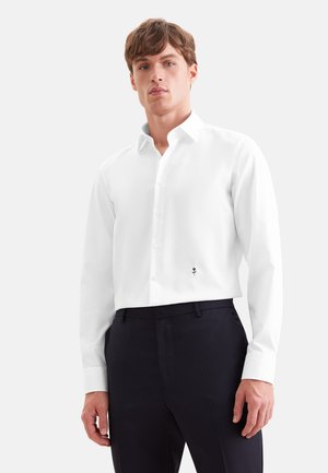 Jeune homme portant une chemise blanche à manches longues avec un petit détail de rose noire près de la taille et un pantalon noir.
