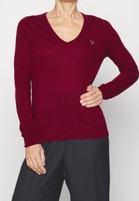 Bordeaux V-Ausschnitt-Pullover aus weichem Strickstoff, mit langen Ärmeln und gerippten Bündchen, sowie einem kleinen Logodetail auf der Brust.