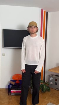 Baltas megztinis su ilgomis rankovėmis, laisvas, derinamas su juodomis tiesiomis kelnėmis. Stovi šalia spalvingos lempos ir knygų.