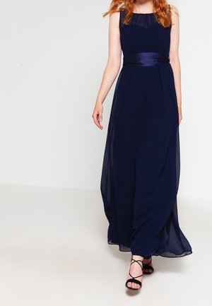 Femme marchant dans une robe longue bleu marine sans manches avec une ceinture en satin et des talons noirs à lanières.