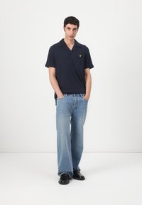 Marinblå polo skjorta med avslappnad krage, korta ärmar och en gul logodetalj. Matchad med ljusblå vida jeans och svarta skor.