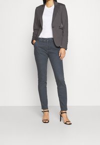 Szara marynarka na dwa guziki, noszona na białej koszulce, zestawiona z ciemnoszarymi dżinsami i czarnymi sandałami na wysokim obcasie z paskami. Gładka tekstura na całej powierzchni.