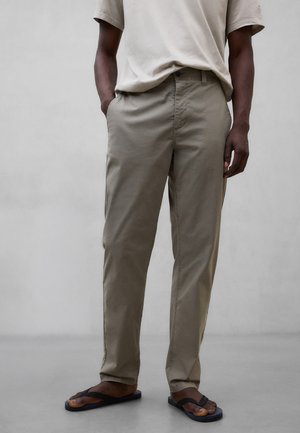 Hombre vestido con pantalones chinos beige, camisa de manga corta beige y sandalias negras, de pie con las manos en los bolsillos contra un fondo gris.