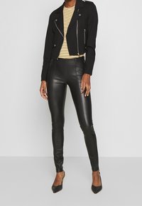 Zwarte cropped leren bikerjack met zilveren ritsen, gecombineerd met een geel en wit gestreept aansluitend shirt en zwarte high-waisted neplederen broek.