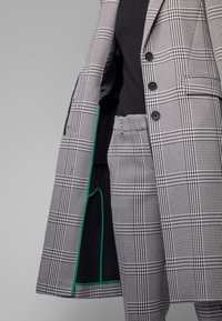 Blazer a cuadros en blanco y negro con un forro acentuado en verde, combinado con pantalones a juego también a cuadros, mostrando detalles de botones y bolsillos.
