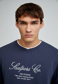 Pulover bleumarin cu guler rotund, cu text alb brodat și o croială lejeră. Textură moale, design minimalist.