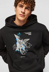 Sudadera negra de algodón, que presenta una impresión gráfica de una figura angelical con acentos azules y un texto en negrita en la parte frontal.