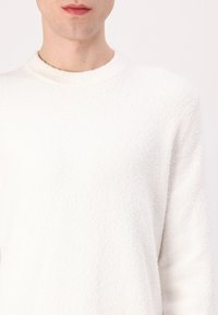 White, textured sweater with a round neckline and long sleeves. The fabric has a soft, fuzzy appearance, offering a cozy look.