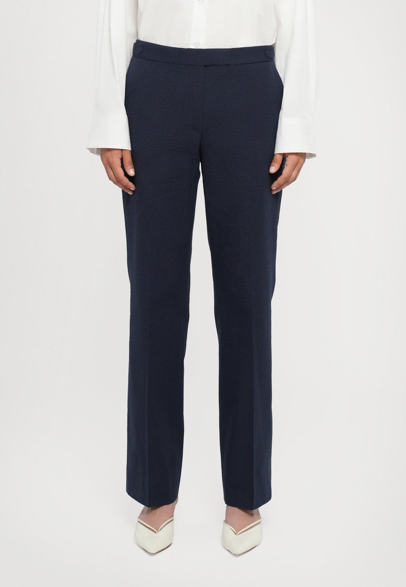 Pantalons ajustés bleu marine en tissu texturé, coupe droite, avec poches avant discrètes. Portés avec une chemise blanche à boutons et des talons beiges.