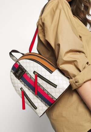 Mochila con un patrón geométrico en blanco, acentuado con rayas rojas, azules y doradas. Incluye tres bolsillos con cremallera y una parte superior de cuero marrón.