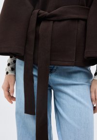 Κοντινό πλάνο ατόμου που φοράει ανοιχτό μπλε τζιν και σκούρο καφέ παλτό με ζώνη πάνω από λευκό τοπ με μαύρες πουά που φαίνονται στους καρπούς.