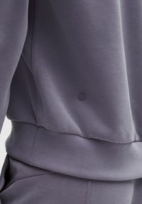 Harmaa urheilupaita; pehmeä, kevyt kangas, jossa on sileä pinta. Siinä on hillitty logo ja resorit helmassa sekä hihojen suissa.