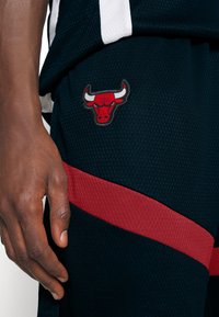 Sorte mesh basketball shorts med en rød vandret stribe. Har et broderet rødt okse-logo. Hånd delvist synlig på shortsene.