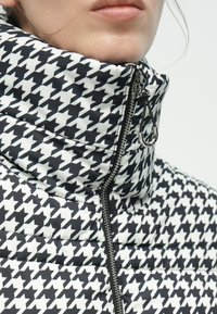 Μπουφάν με επένδυση και ψηλό γιακά, με ασπρόμαυρο μοτίβο ψαροκόκκαλο. Κλείσιμο με φερμουάρ και ασημένιο διακοσμητικό τραβηχτικό.