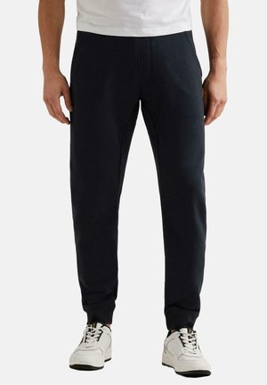 Man die zwarte joggerbroeken en witte sneakers draagt, staand tegen een effen witte achtergrond.