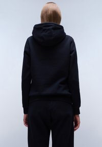 Sudadera negra con capucha, de corte relajado, textura suave y puños acanalados, mostrada desde la parte posterior sobre un fondo gris claro.