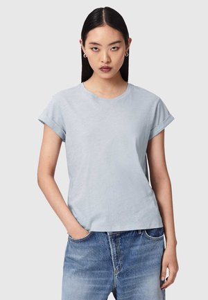 Ung kvinde med glat mørkt hår iført en lyseblå t-shirt med opkørte ærmer og blå jeans, står med den ene hånd i lommen.
