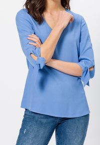 Blouse bleu clair avec un col en V, des manches trois-quarts avec des accents noués, et une texture lisse, associée à un jean bleu.