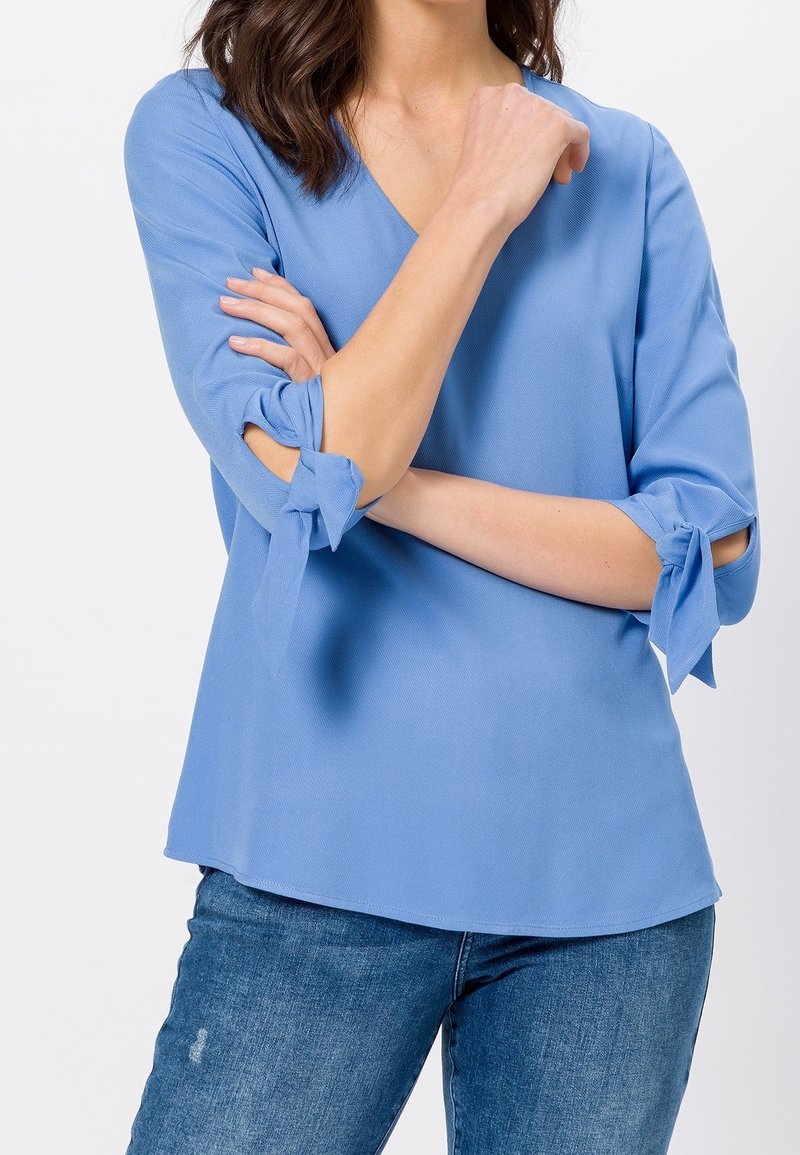 Blouse bleu clair avec un col en V, des manches trois-quarts avec des accents noués, et une texture lisse, associée à un jean bleu.