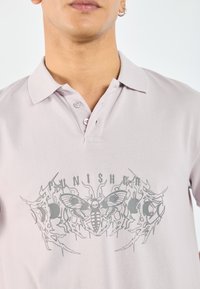 Lyse lilla polo skjorte med krage. Fronten har et grått grafisk design med et hodeskalle og en møll, omkranset av skarpe detaljer.