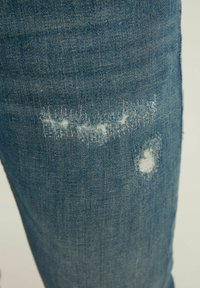 Jambe de jean en denim bleu avec deux déchirures usées laissant apparaître des fils blancs, sur un fond clair uni.