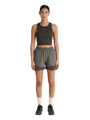 Jonge vrouw met een zwarte mouwloze cropped top met de tekst "HALO", grijze sportshorts, witte sokken en zwarte sportschoenen, rechtopstaand.