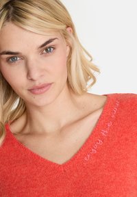 Korallenroter V-Ausschnitt-Pullover mit weichem Material. Mit dem gestickten Satz "genieße den Moment" in Pink in der Nähe des Ausschnitts.